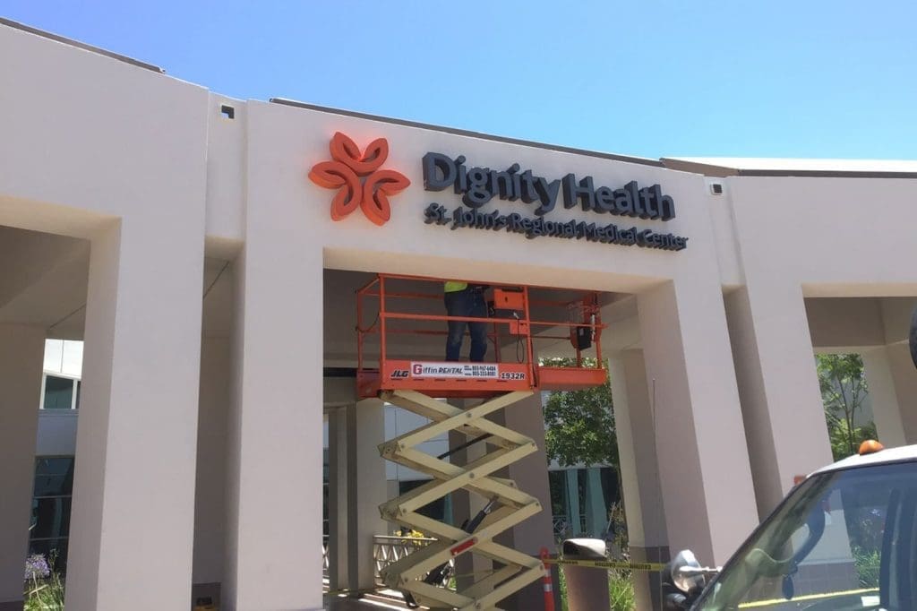 A worker on a scissor lift installs or repairs the Dignity Health St. Louise Regional Medical Center sign on a building exterior.