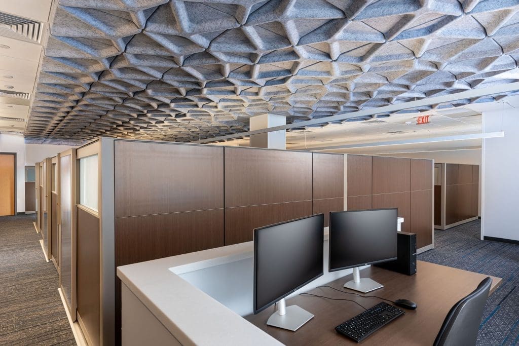 Modern office space with wood-paneled cubicles, dual computer monitors on a desk, and a geometric acoustic ceiling design.