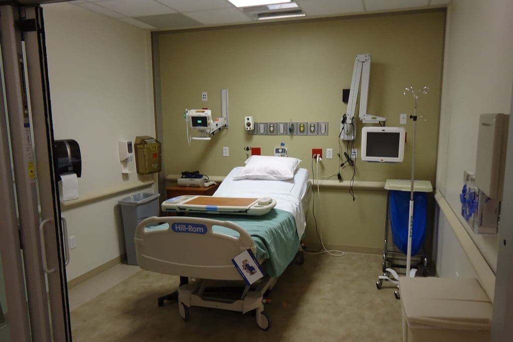 A hospital room with a bed, medical equipment, monitor, and supplies against a beige wall, viewed from the entrance.