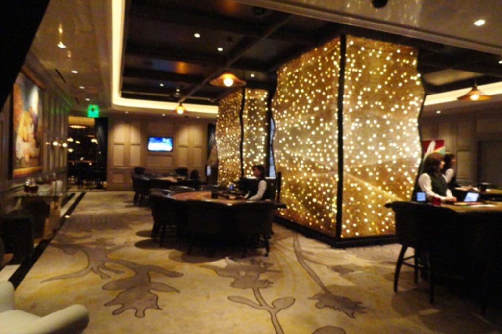A dimly lit casino interior with two large, illuminated panels, several gaming tables, and staff in uniforms.