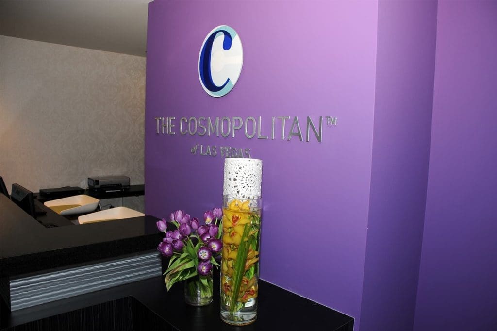 Reception desk at The Cosmopolitan of Las Vegas with a purple wall, company logo, fresh flowers, and a decorative vase.