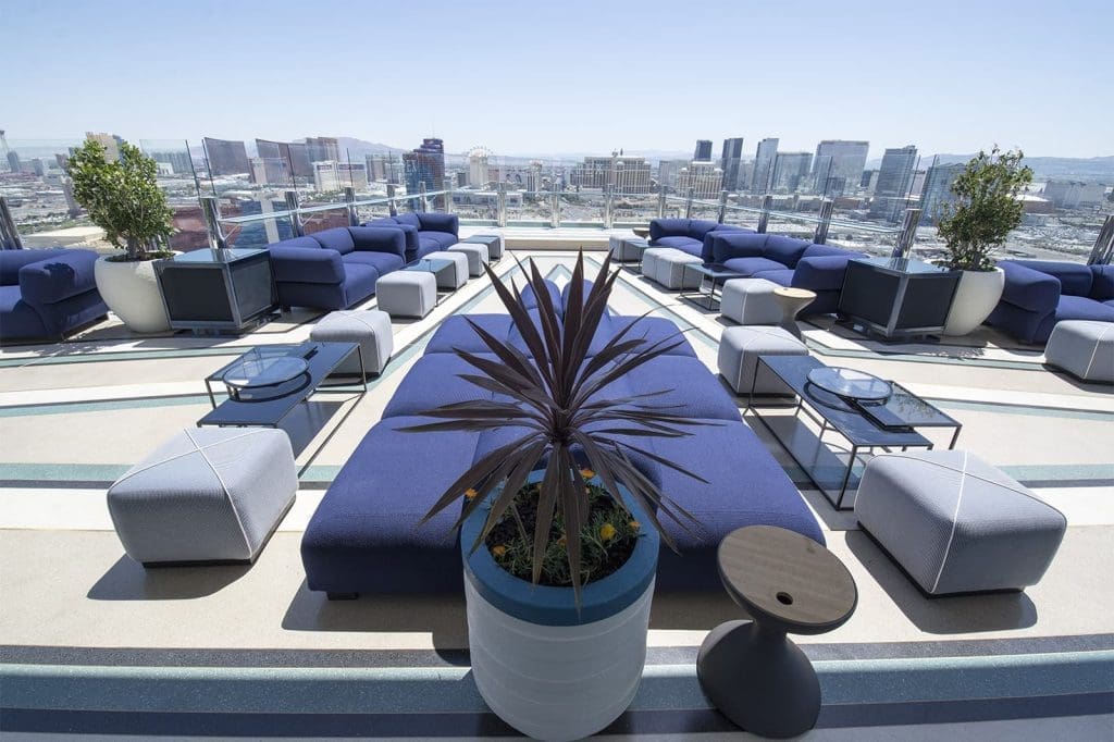 Rooftop lounge area with blue cushioned seating, tables, potted plants, and a city skyline visible in the background under a clear sky.