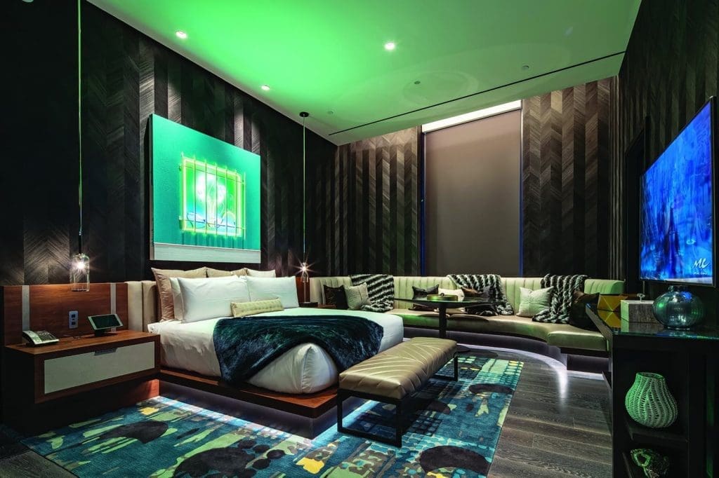 Modern hotel room with a large bed, sectional sofa, illuminated artwork, patterned rugs, and green accent lighting, creating a contemporary and cozy atmosphere.