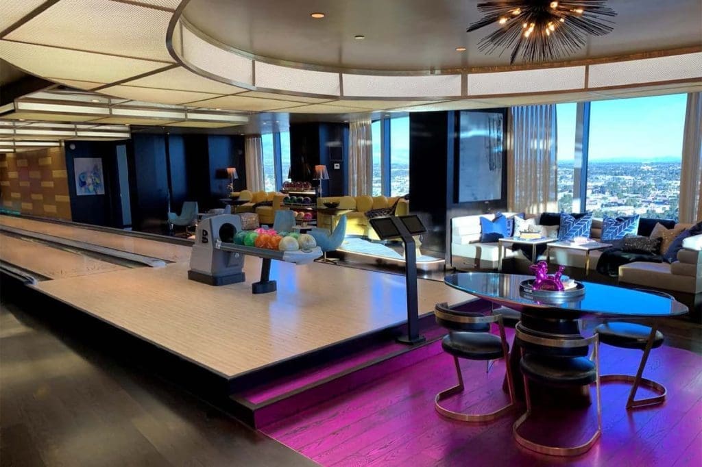 Modern home interior featuring a private bowling alley, colorful bowling balls, a round dining table with chairs on a magenta rug, and large windows with a city view.