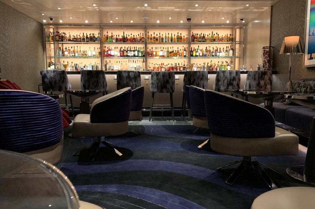 Modern bar interior with blue and gray chairs, high tables, and a well-lit back wall displaying various bottles of alcohol.