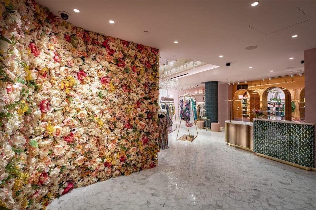 Interior of a boutique with a wall covered in artificial flowers, marble flooring, clothing racks, and a modern, stylish decor.