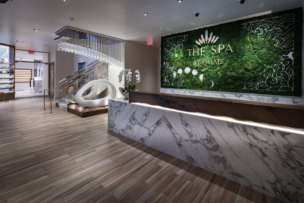 Modern spa reception area with marble front desk, large green wall art labeled "The Spa at Palms," wood flooring, and a sculptural white seating area near a staircase.
