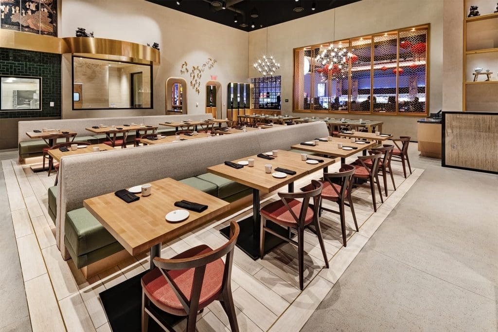 Modern restaurant interior with wooden tables, red and green chairs, booth seating, wall art, chandeliers, and place settings neatly arranged.