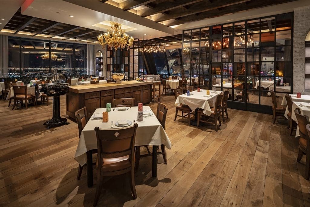 Elegant restaurant interior with wooden floors, white tablecloths, chandelier lighting, large windows, and city views in the background.