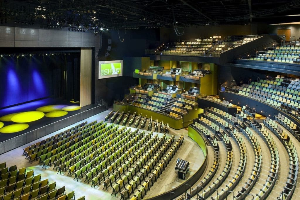 A large, modern theater auditorium with rows of empty seats, a stage lit with yellow spotlights, and a screen displaying the word "SHOW.