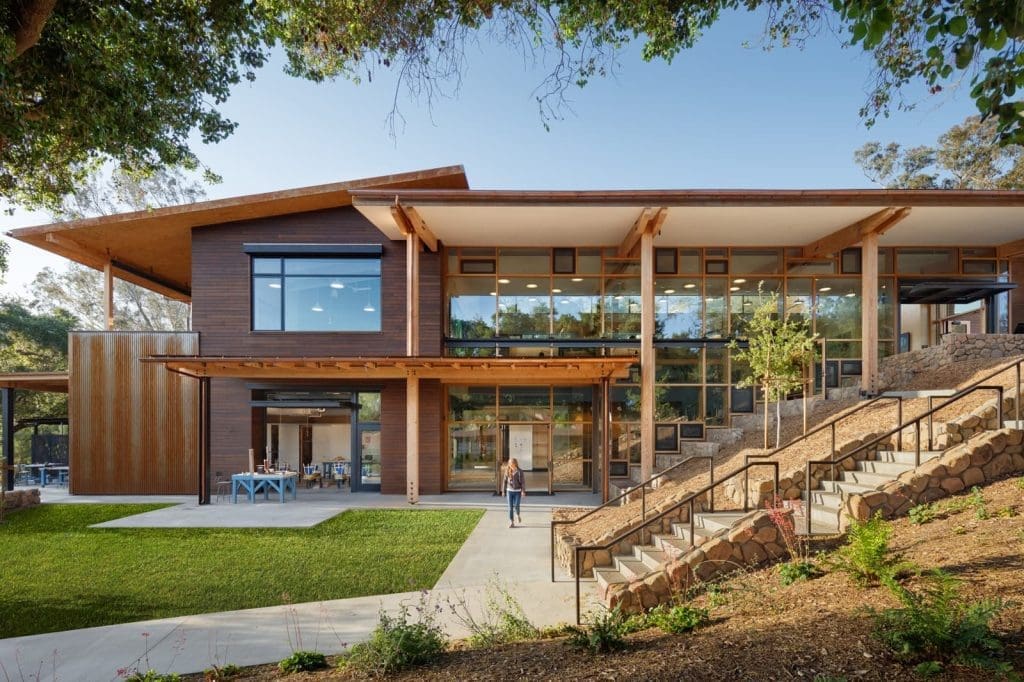 A modern two-story building with large windows, wooden accents, and a sloped roof, surrounded by greenery and a stone staircase.
