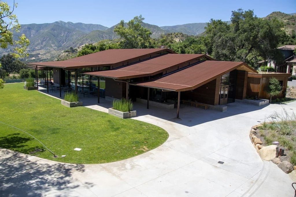 Modern building with angled metal roofs, large glass walls, and wooden exterior sits on a landscaped lawn with mountains and trees in the background.