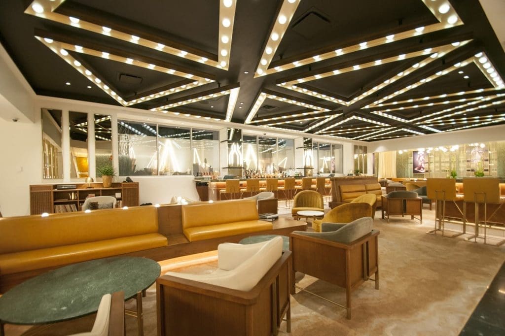 Modern lounge with geometric ceiling lights, brown leather sofas, armchairs, and tables arranged in a spacious, well-lit area with a mirrored wall and decorative plants.