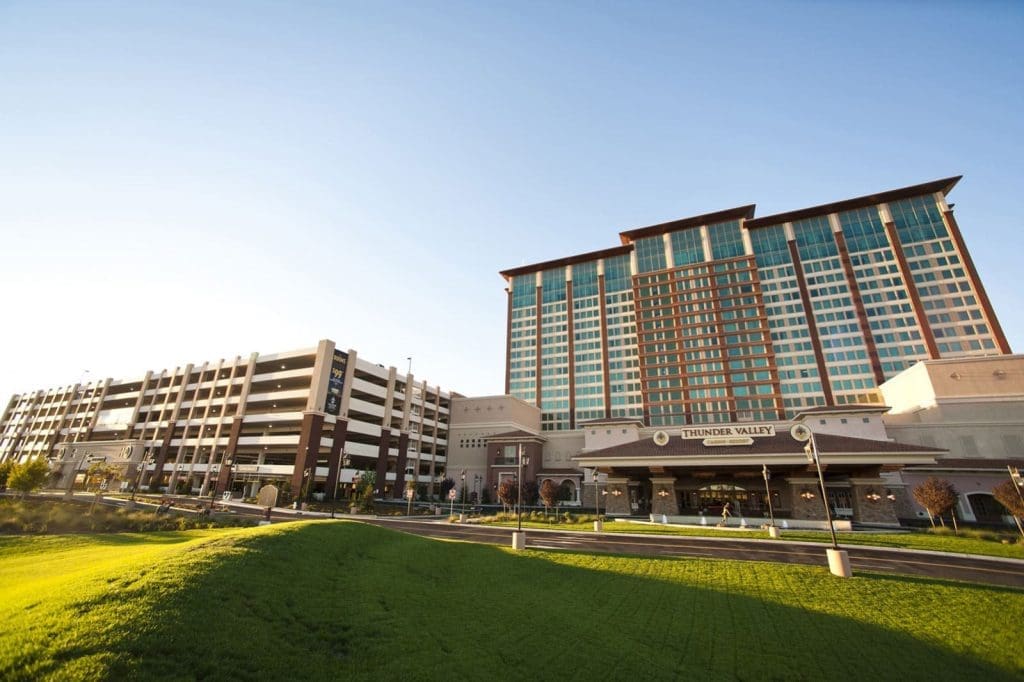 A large hotel and casino building with "Thunder Valley" signage stands next to a multi-level parking structure under a clear sky, with green grass in the foreground.