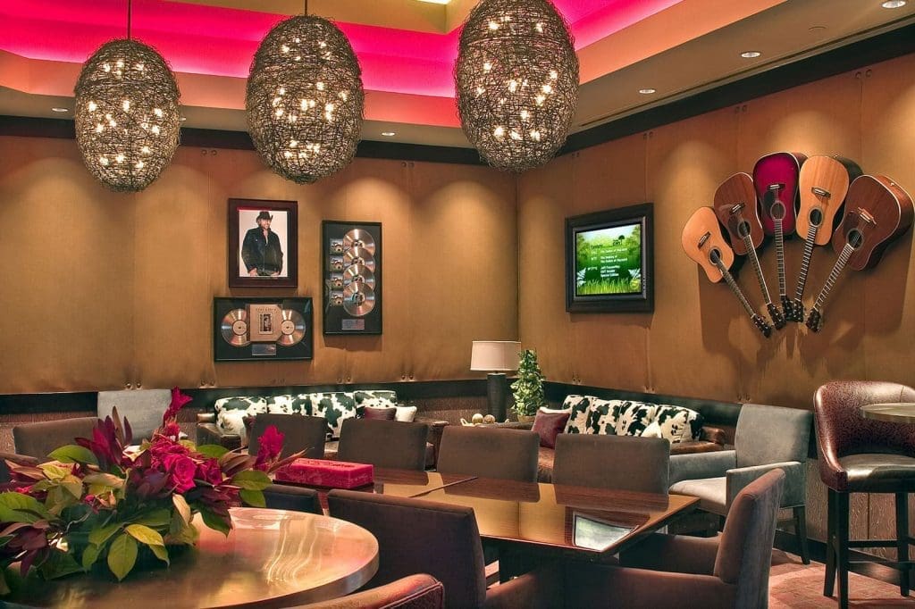 A lounge with modern lighting, wall-mounted guitars, framed music memorabilia, a TV, assorted chairs and tables, and a floral centerpiece on one table.