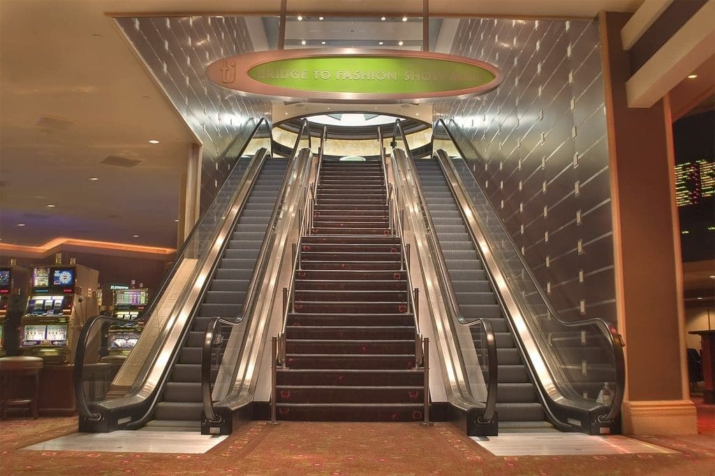 A set of three escalators, two going up and one going down, with a sign above reading "BRIDGE TO FASHION SHOW MALL" inside a casino.