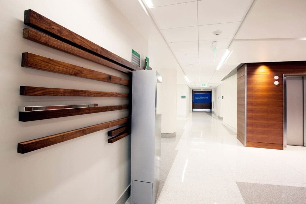 Modern hospital hallway with light walls, polished floors, wooden accents, and an elevator on the right. A decorative wood panel and sanitizer dispenser are mounted on the left wall.