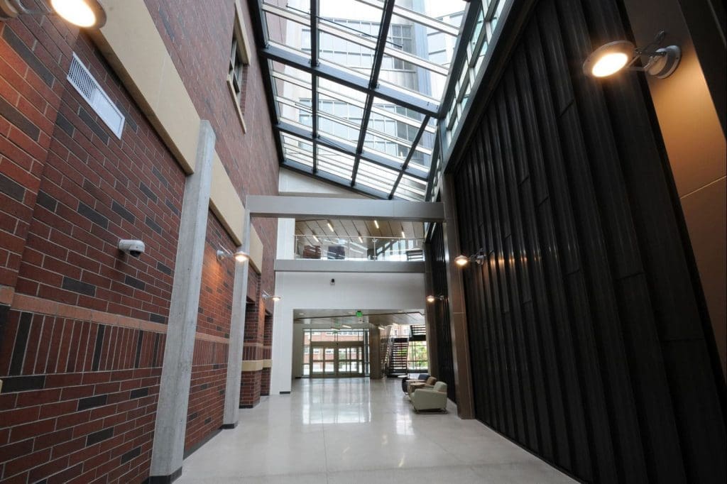 A modern building lobby with brick and dark panel walls, glass ceiling, overhead lights, and a seating area near glass entrance doors.