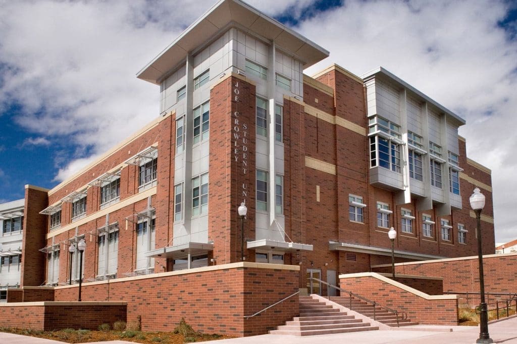 A modern brick and glass building with large windows, multiple stories, and stairs leading to the entrance, set against a partly cloudy sky.