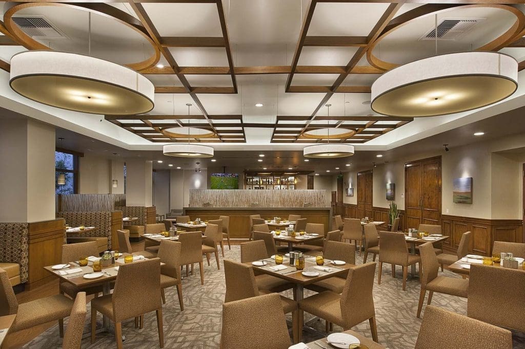 A modern restaurant dining area with wooden tables, cushioned chairs, neutral decor, and large circular ceiling lights. Tables are set with plates, cups, and condiments.
