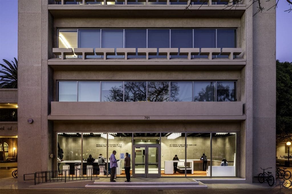 A modern building with large windows and a well-lit entrance; several people are entering and sitting at desks inside near the glass facade.