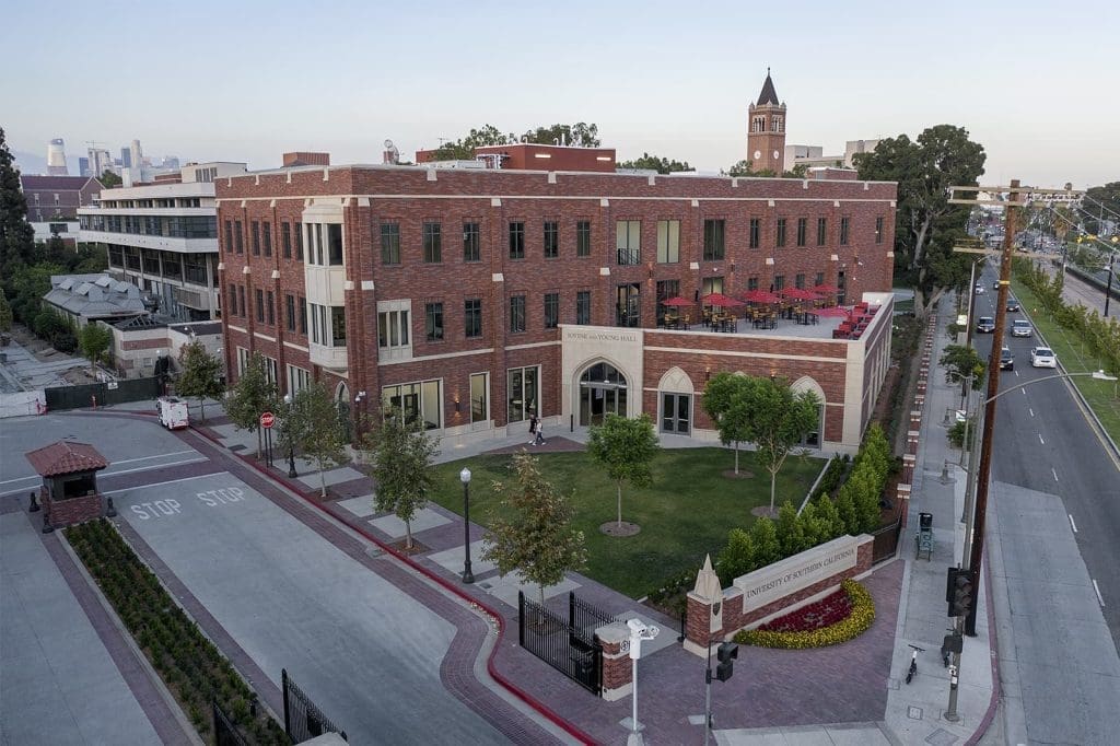 Aerial view of a multi-story red-brick building on a neatly landscaped campus with a clock tower in the background and surrounding roads visible.