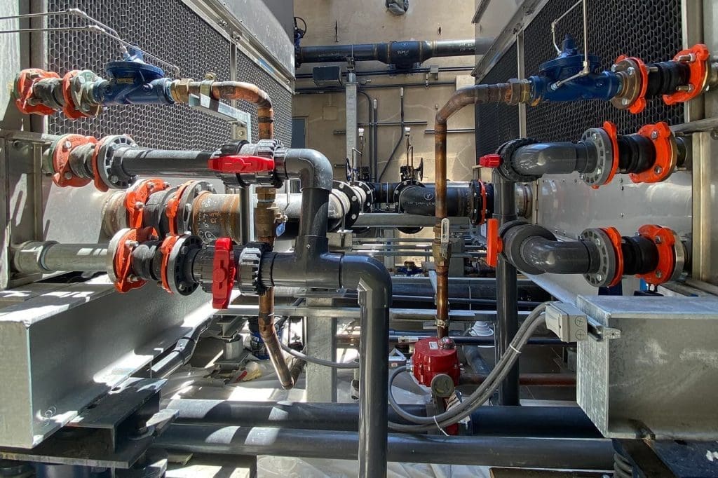 Industrial piping system with multiple valves, connectors, and metal pipes, featuring red and blue valve handles, set between two large units.