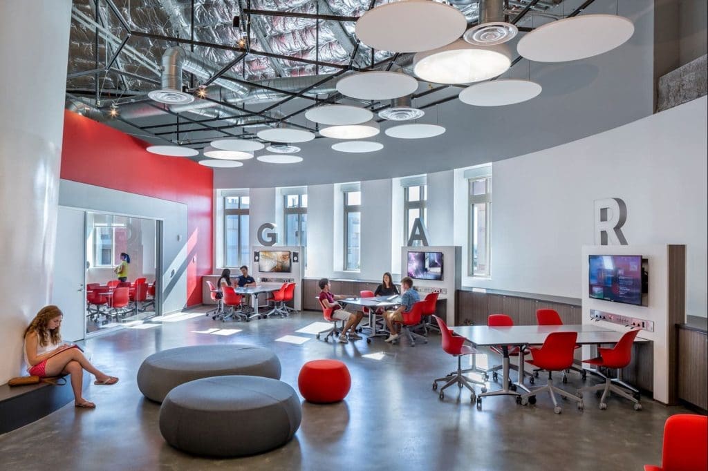 Modern classroom with red chairs, round gray seating, wall-mounted monitors, and students working in groups; exposed ceiling and large letters on walls.