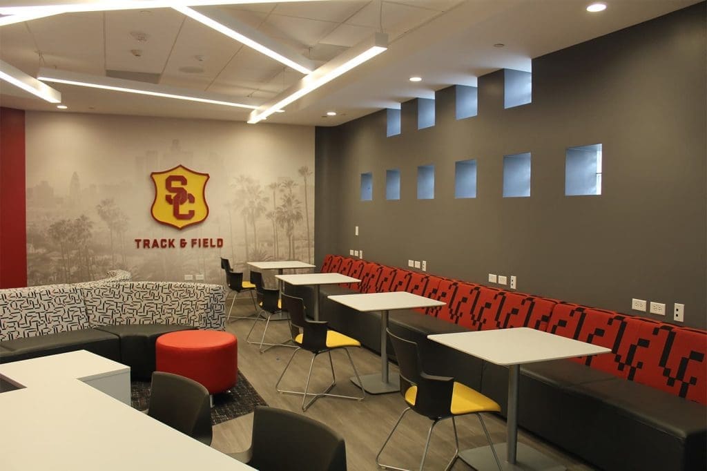Modern lounge area with tables, chairs, and a long bench featuring USC Track & Field branding on the wall and square windows on a gray accent wall.