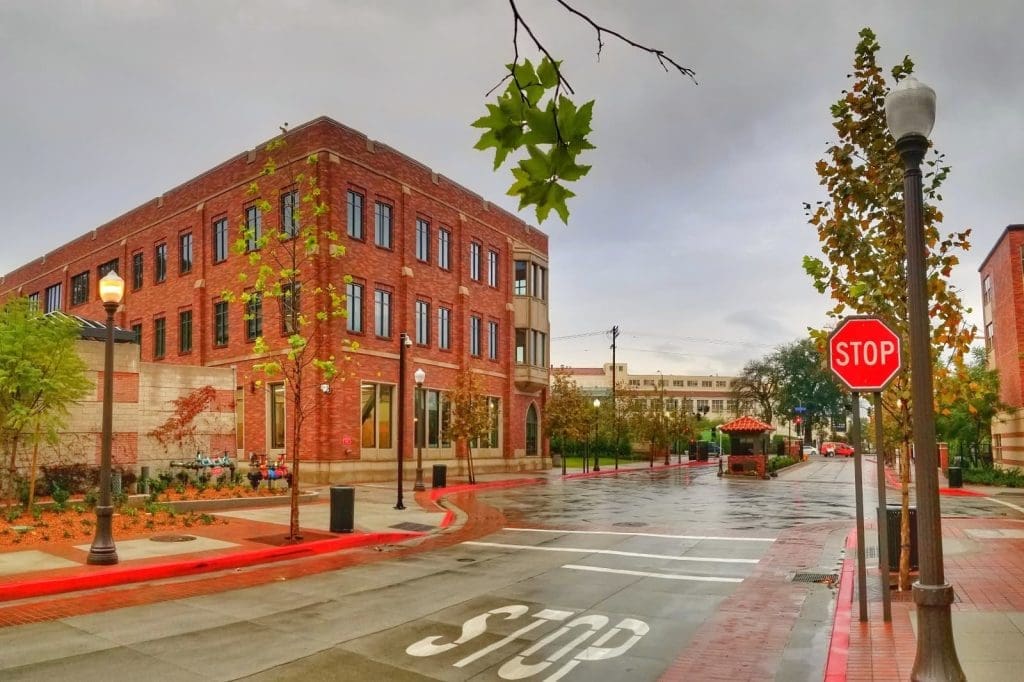 A wet, empty street corner with a red brick building, stop signs, street lamps, and scattered trees under an overcast sky.