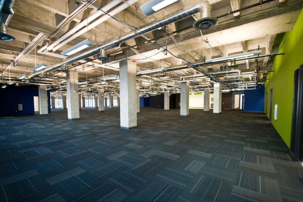 Large empty office space with exposed concrete ceiling, visible ductwork, carpeted floor, and pillars. One wall is painted green, the others are dark blue. Natural light enters through windows.