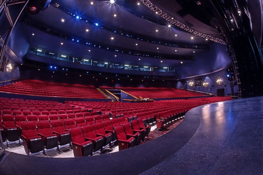 Photo of an empty theater auditorium with multiple tiers of red seats, seen from the stage under blue lighting.
