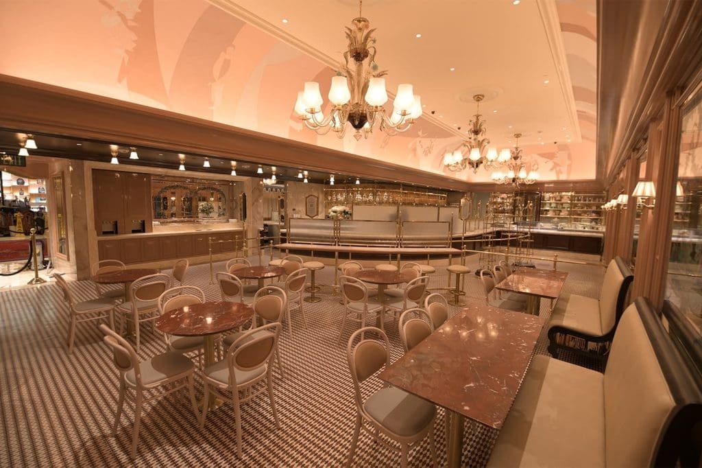 Spacious cafe with round marble tables, beige chairs, chandeliers, and a patterned tile floor under warm lighting. The bar and shelves line the back wall.