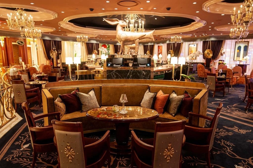 Elegant restaurant interior with plush seating, ornate chandeliers, decorative pillows, a central marble counter, and a large gold dachshund statue above the bar.