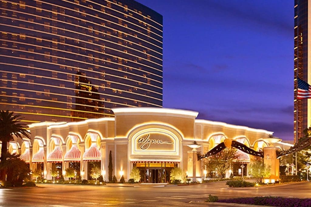 The illuminated entrance of the Wynn hotel and casino in Las Vegas at dusk, with a modern high-rise building and palm trees in the background.