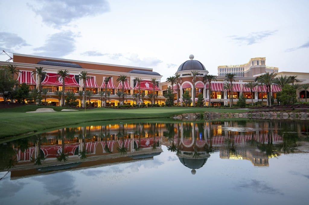 A large building with red awnings and palm trees is reflected in a pond at dusk, with some clouds visible in the sky.