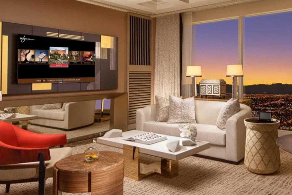 A modern hotel living room features a large TV, neutral-toned furniture, a coffee table, and a large window with a view of a cityscape at sunset.