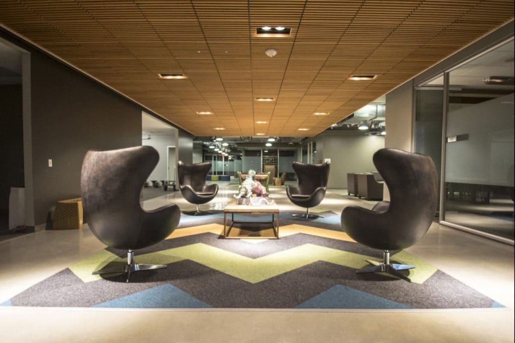 Modern office lounge with four black egg chairs around a coffee table on a chevron-patterned rug, under a wooden panel ceiling with glass walls in the background.