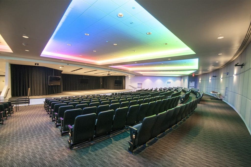 Empty auditorium with rows of black seats, a stage with black curtains, large display screens, and ceiling lights in changing colors.
