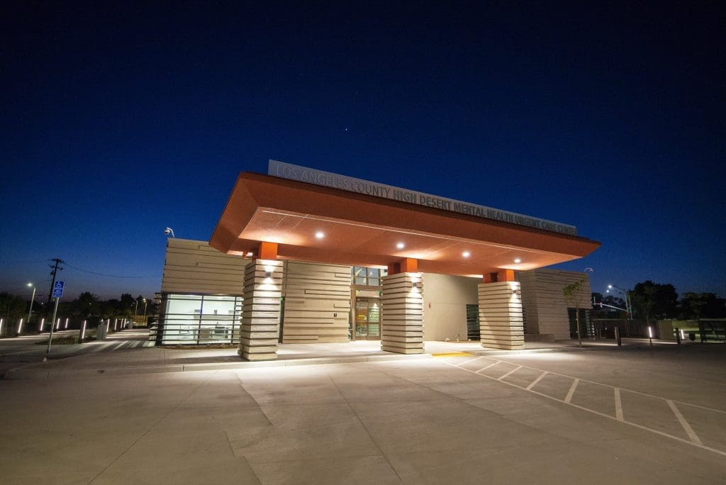 A modern building with an illuminated entrance at dusk, labeled "Los Angeles County High Desert Mental Health Urgent Care." The parking lot in front appears empty.