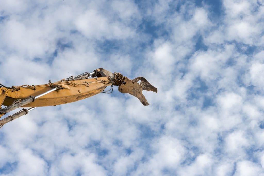 An excavator arm with a metal attachment is extended upward against a background of partly cloudy sky.