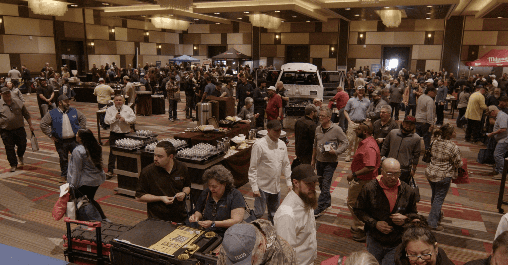 Large group of people attending an indoor event or expo, with booths, tables, and displays set up in a spacious, well-lit hall.