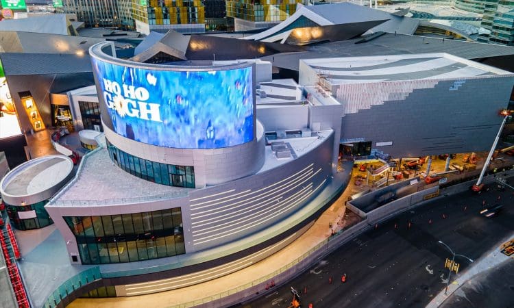 Aerial view of a modern commercial building with a large curved digital screen displaying "HO HO" above a city street with traffic cones and ongoing construction.
