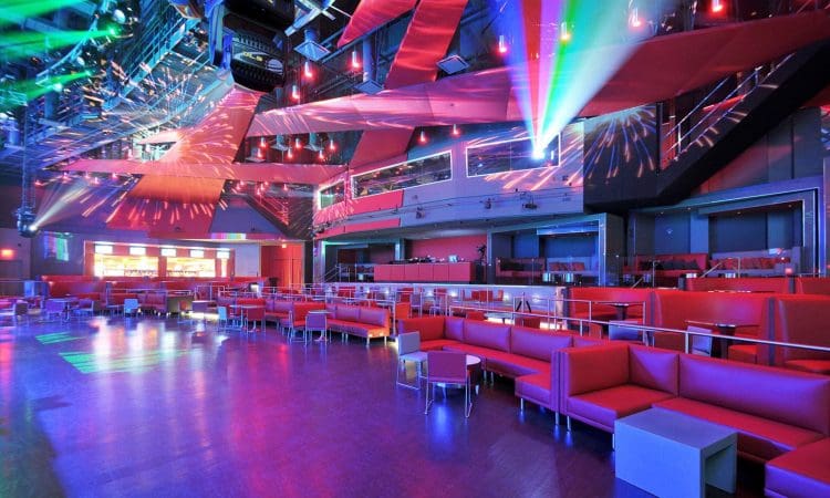 Large modern nightclub interior with red seating, colored lights, reflective ceiling, and spacious dance floor.