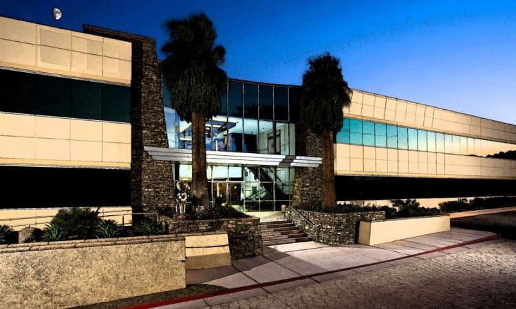 Modern two-story office building with large glass entrance, stone accents, palm trees, and night sky in the background.