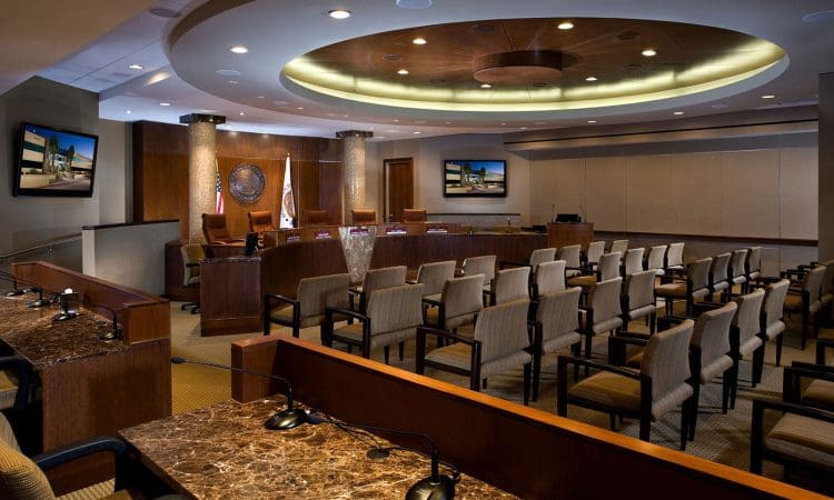 Modern council chamber with rows of beige chairs, microphones, a marble desk, large screens, and a circular ceiling with recessed lighting. No people present.