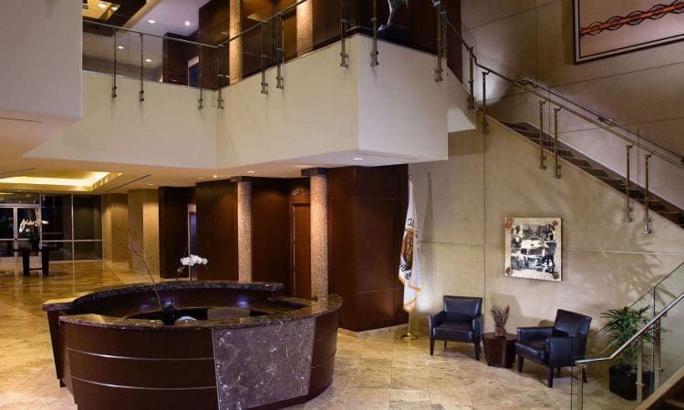 Modern office lobby with a round marble reception desk, two black chairs, a staircase, and decorative artwork on beige walls.