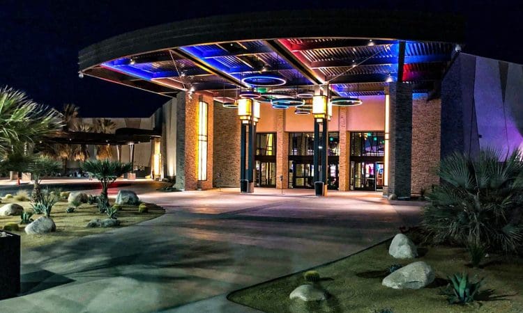 The entrance to a modern building at night, featuring colorful overhead lights, large glass doors, and landscaped desert plants.