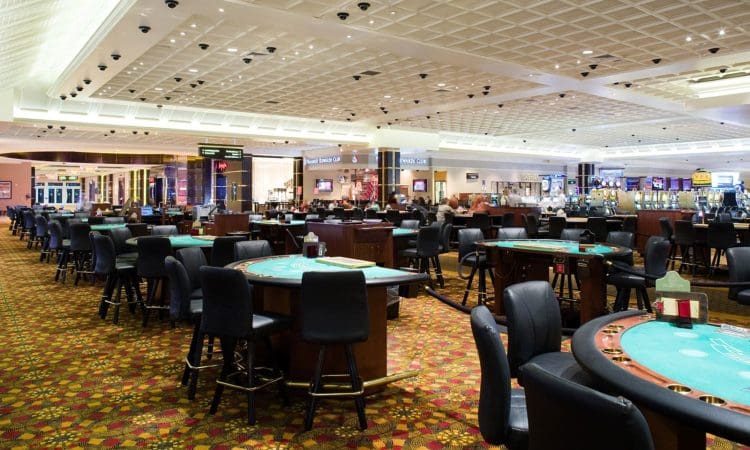 A spacious casino floor with empty gaming tables and chairs, patterned carpet, and bright overhead lighting. Slot machines and signs are visible in the background.