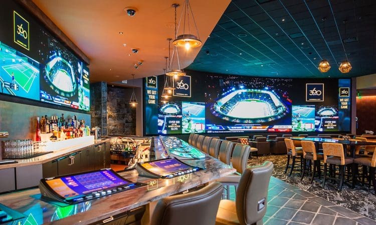 Modern sports bar with large screens displaying sporting events, a long bar with stools, and a row of betting kiosks on the counter.
